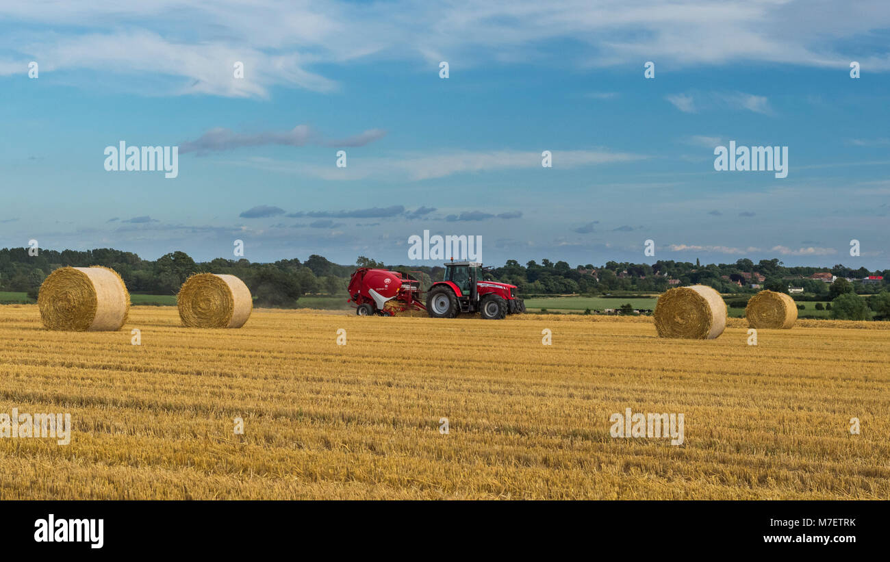 Farmer in red farm tractor pulling round baler, is driving & working in field, baling straw into round bales - Whixley, North Yorkshire, England, UK. Stock Photo