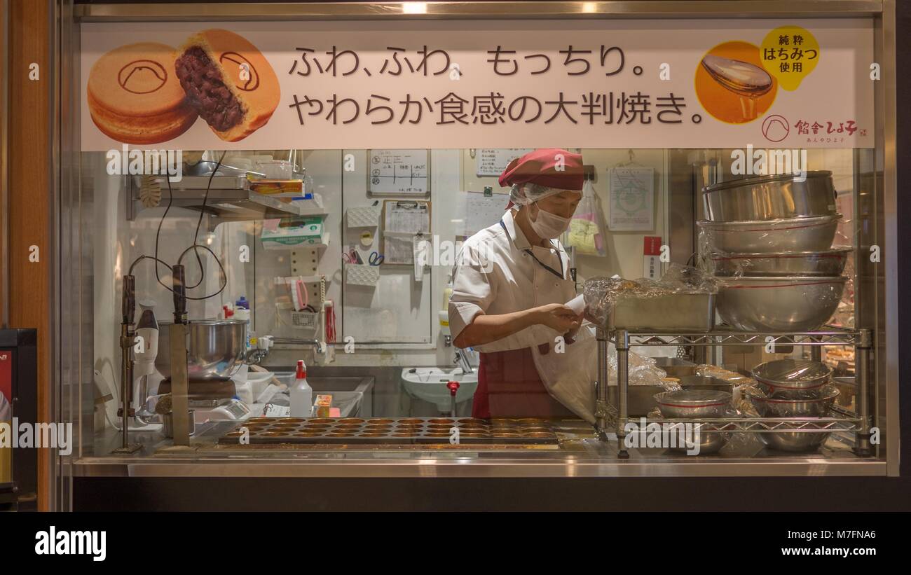 Japanese baker wearing red hat and apron surrounded by bowls making traditional Japanese red bean cakes shot through bakery window. Stock Photo