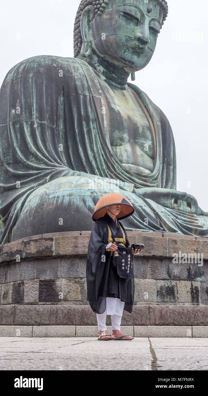 Japanese Buddhist pilgrim in straw hat, black robes and white pants chanting sutras, ringing bell and holding black lacquered bell dwarfed by Buddha Stock Photo