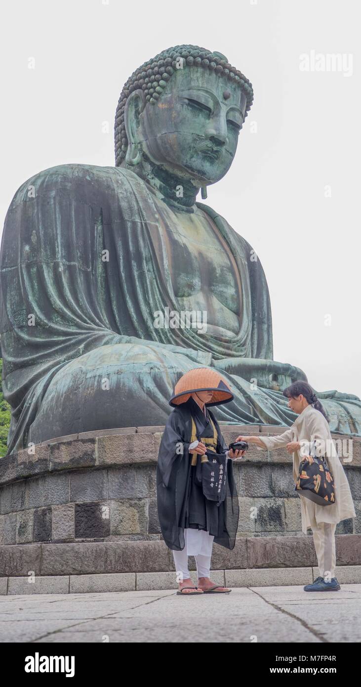 Japanese woman placing money in black lacquered bowl of Japanese Buddhist pilgrim in front of Kamakura Buddha ringing bell and chanting sutras. Stock Photo