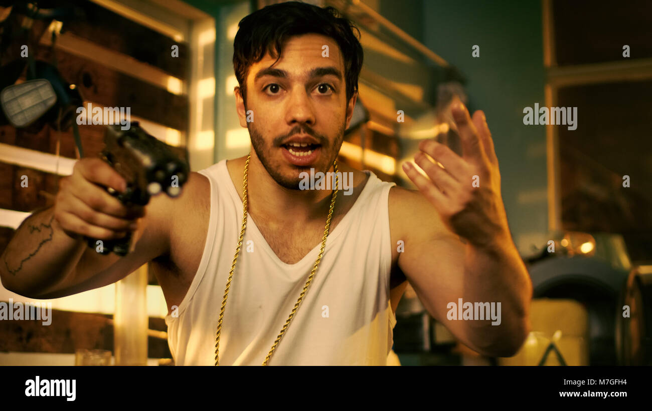 Brutal Gang Member Gone Crazy Makes Threatening Gestures with a Gun. He Wears Gold Chain and Sleeveless Shirt. In the Background Abandoned Building. Stock Photo