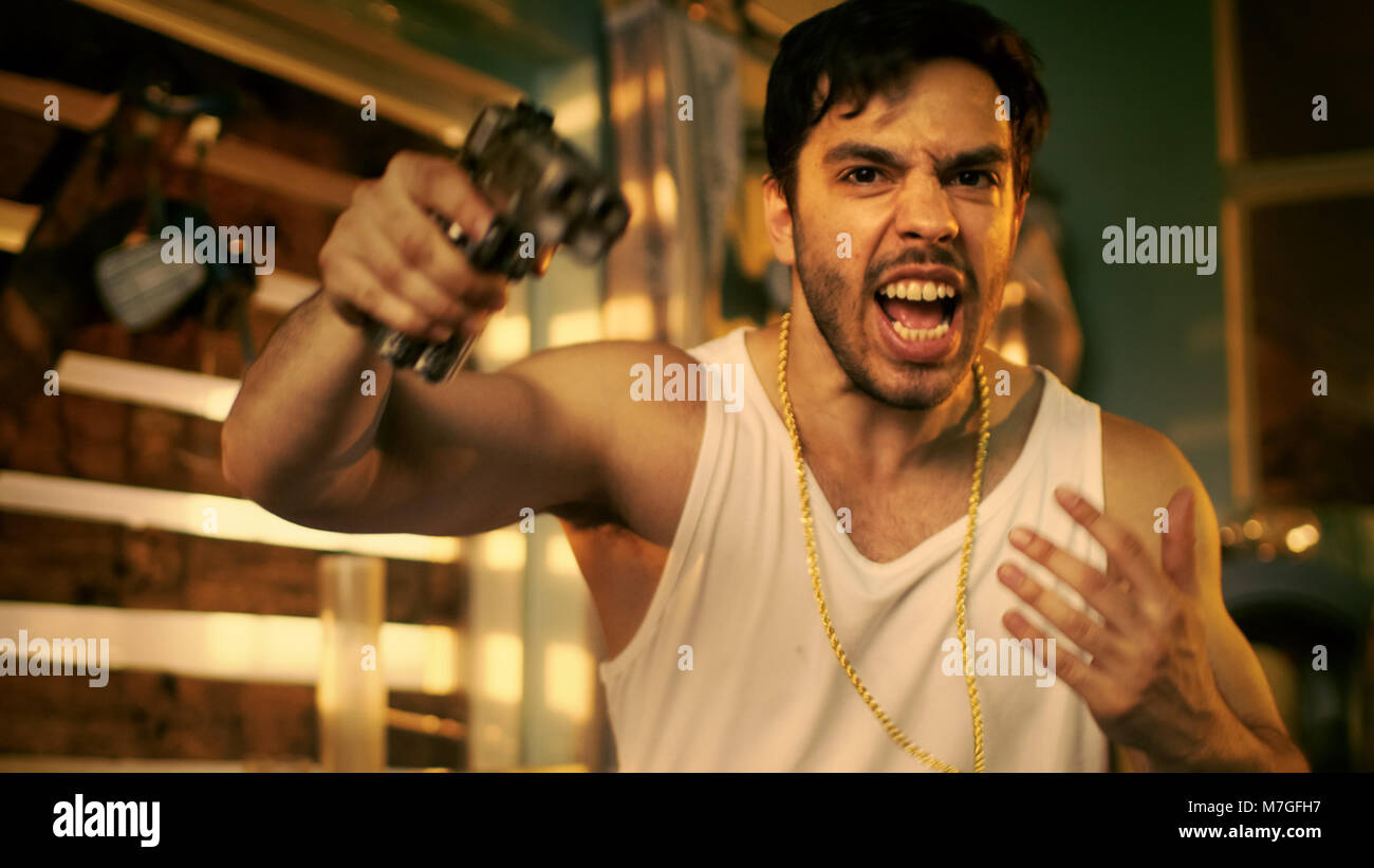 Brutal Gang Member Gone Crazy Makes Threatening Gestures with a Gun. He Wears Gold Chain and Sleeveless Shirt. In the Background Abandoned Building. Stock Photo