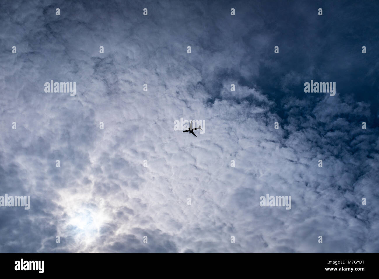 Airplane in silhouette flying across textured cirrocumulus clouds seen from the ground Stock Photo