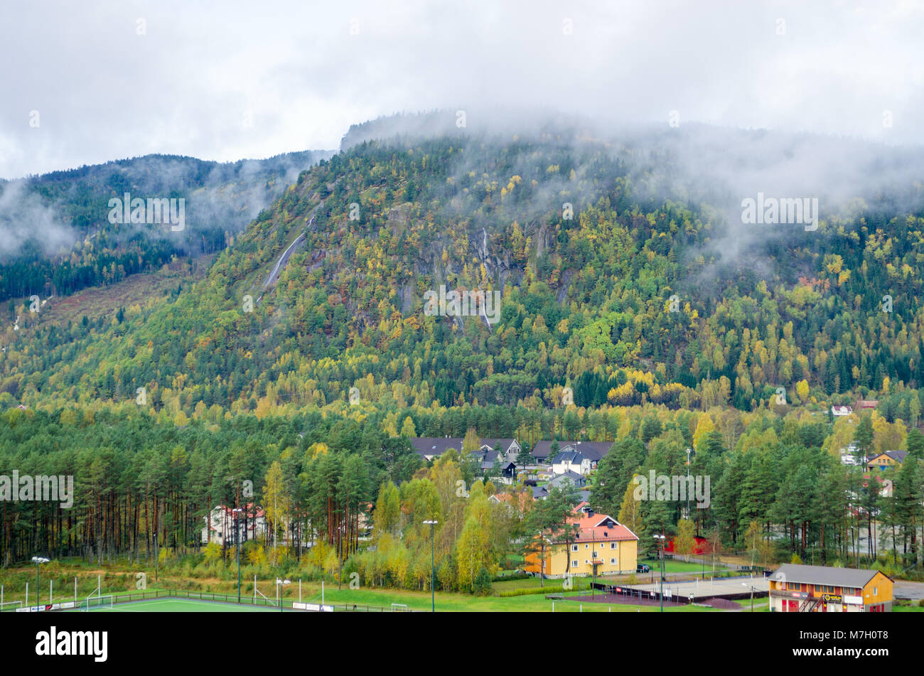 Long distance view of Evje cityline dominated by low mountain covered by forest and clouds, central Norway, Europe. Stock Photo