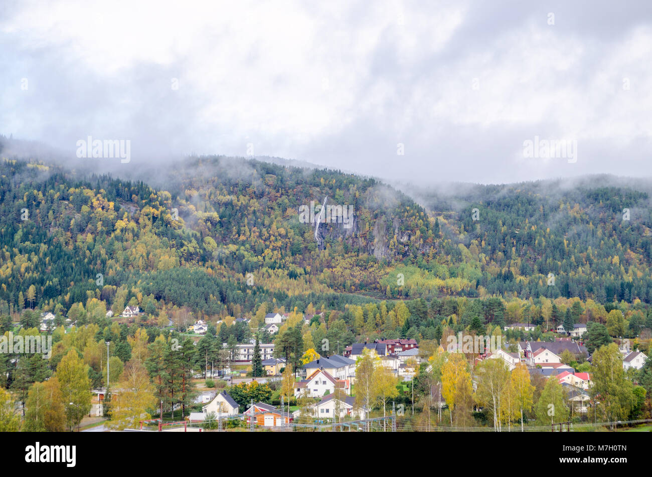Long distance view of Evje cityline dominated by low mountain covered by forest and clouds, central Norway, Europe. Stock Photo