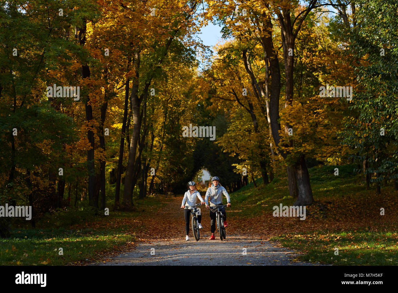 Happy smiling young couple wearing healmets and sportswear going for a bike ride on a sunny warm autumn day in the park. They are having fun together. Stock Photo