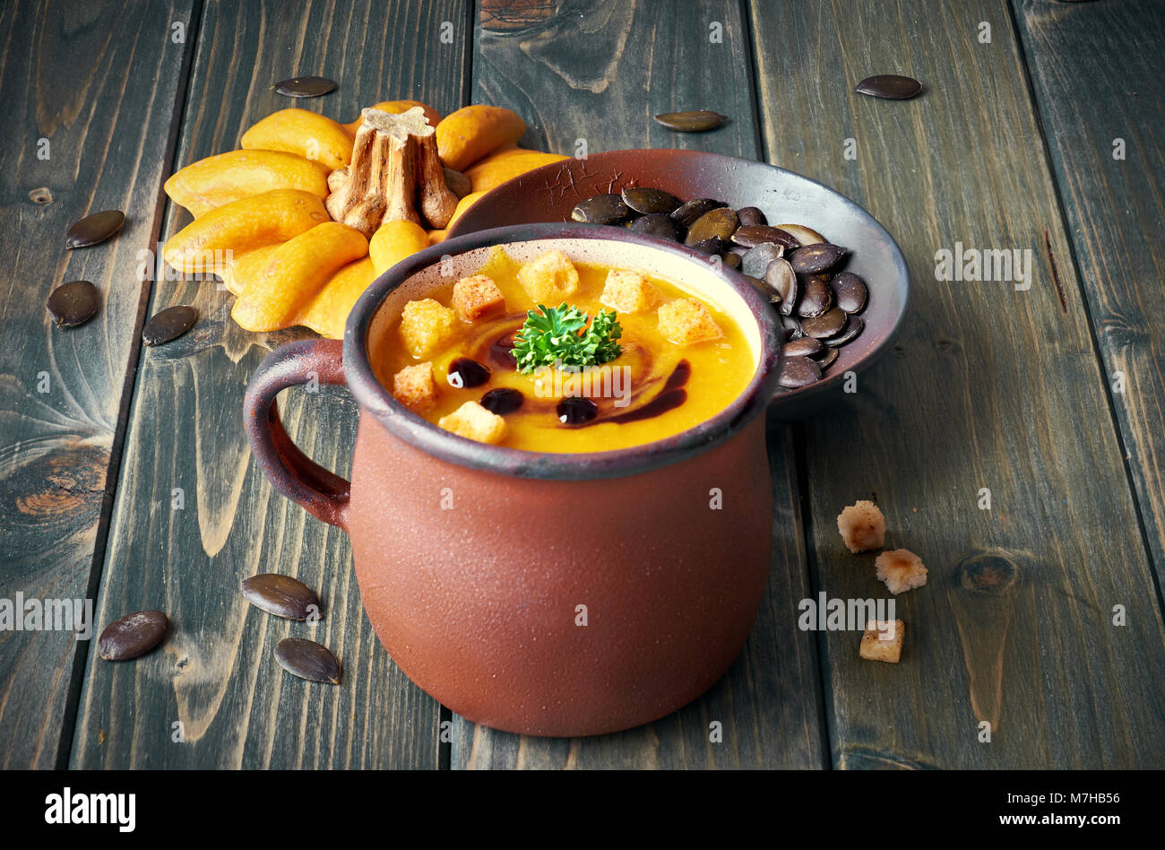 Tasty pumpkin soup in old enamel mug on rustic wooden table. The soup is served with croutons, parsley and pumpkin oil. Stock Photo