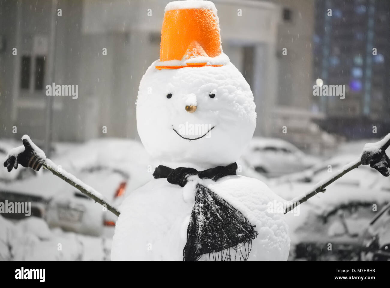 Snowman on a blurred background in gray with blue lights of the cityscape. Closeup of smiling snowman with orange hat, scarf and carrot nose, outdoors Stock Photo
