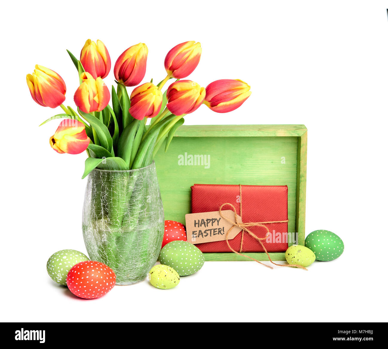 Bunch of red tulips with yellow tops, wooden board, gift with tag and colored Easter eggs on white, text 'Happy Easter' on the tag Stock Photo