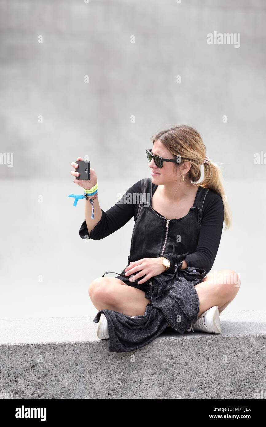 Young blonde lady with Ponytail, sitting on wall with mobile phone, taking a selfie, Bilbao, Vizcaya, Pais Vasco, Spain, Stock Photo