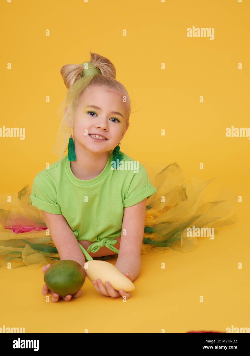 The fair-haired little girl wears a light green t-shirt and a bright colorful skirt.Holding the baby holding the mango fruits of different varieties.  Stock Photo