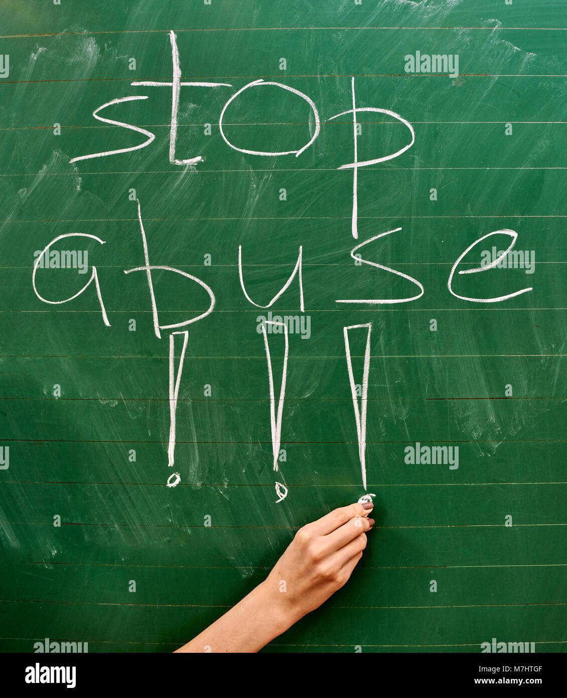 Right hand of a young girl writing the words STOP ABUSE on the green school board with a white chalk vertical background Stock Photo