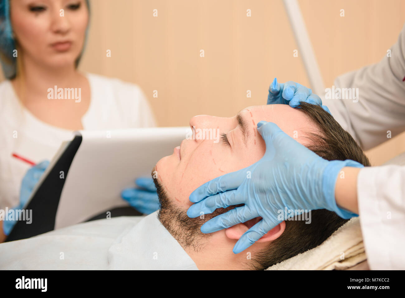 Consultance for man in private clinic of plastic surgery. Two doctors and patient. Hands of doctors in rubber blue gloves touch face. Stock Photo