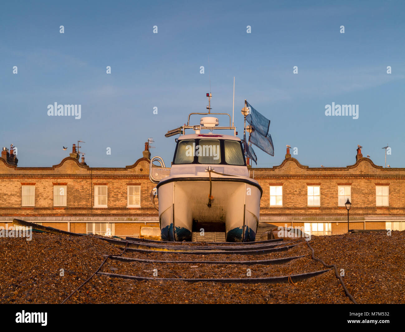 Front on symmetrical shot of a twin hull fishing boat moored on a bank of shingle in front of a building facade with evening light Stock Photo