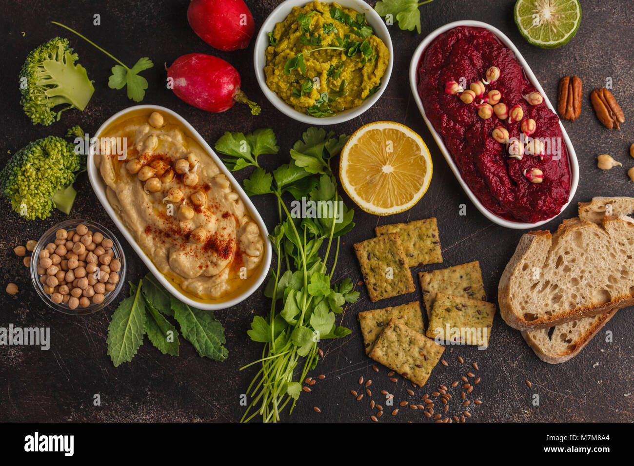 Vegan food background. Vegetarian snacks: hummus, beetroot hummus, green peas dip, vegetables, tofu. Top view, dark background, copy space. Stock Photo