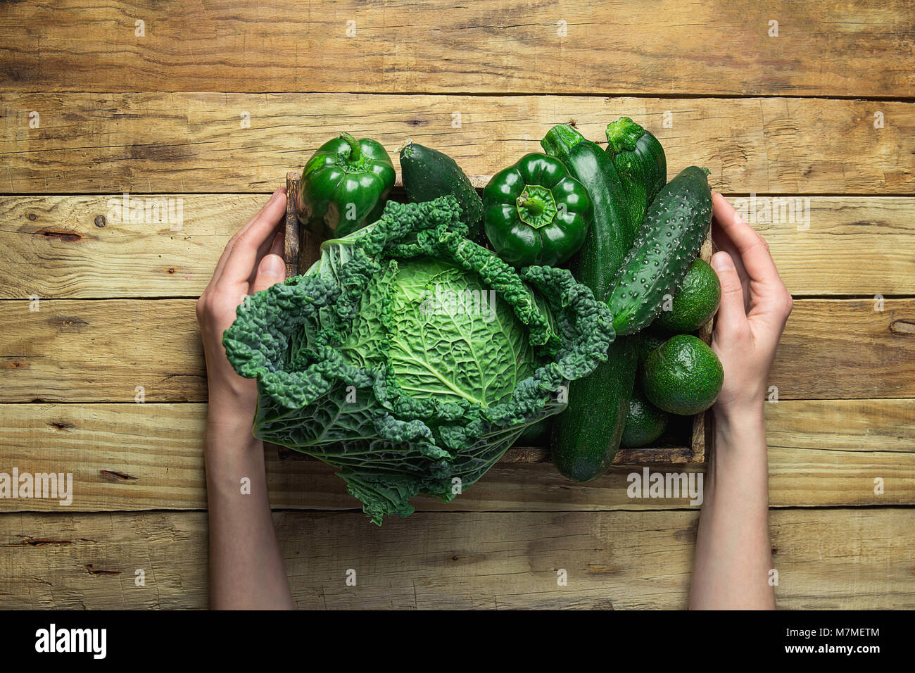 Young Woman Holding in Hand Trey with Fresh Organic Green Vegetables Savoy Cabbage Zucchini Cucumbers Bell Peppers Avocados on Aged Plank Wood Backgro Stock Photo