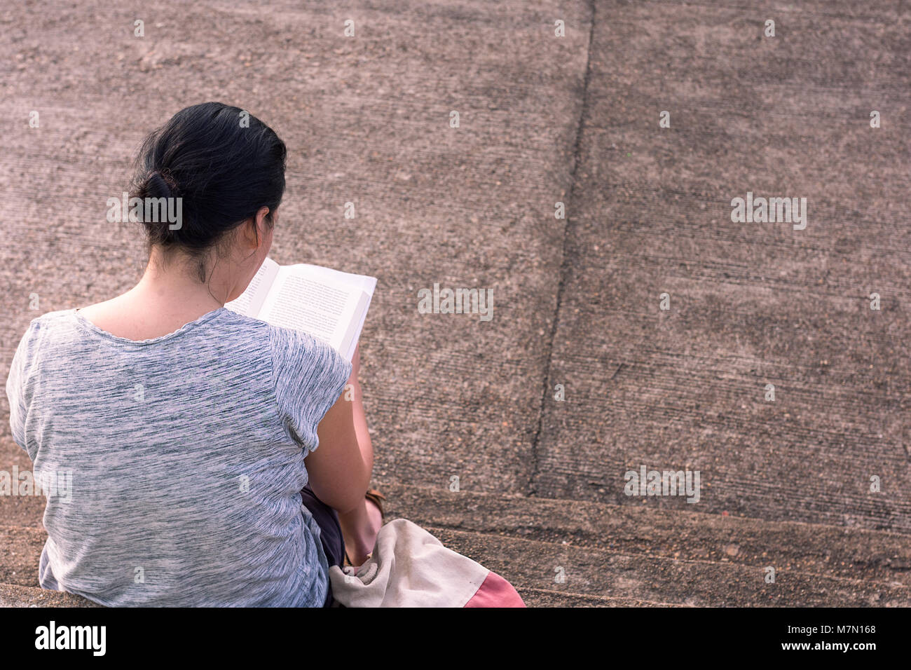 A young woman concentrates reading here hard back book whilst sat on some concrete steps. Stock Photo