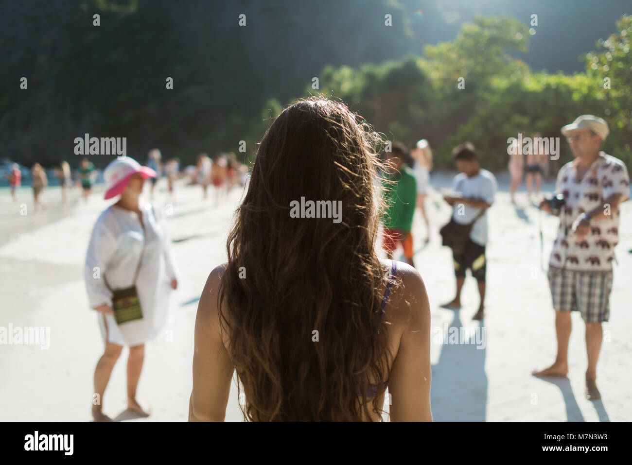 Back view of female traveler is standing on the beach with group of tourists. Young unrecognizable woman under the sun rays. Blurred people Stock Photo