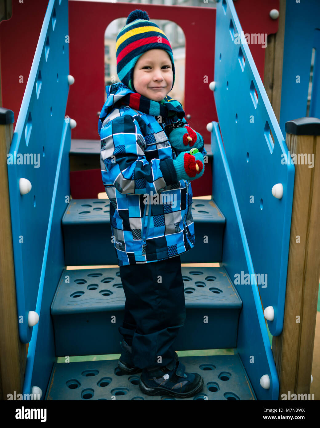Cheerful little boy is standing on the staircase at the playground and holding toys in hands. Beautiful child smiling and playing in the yard Stock Photo