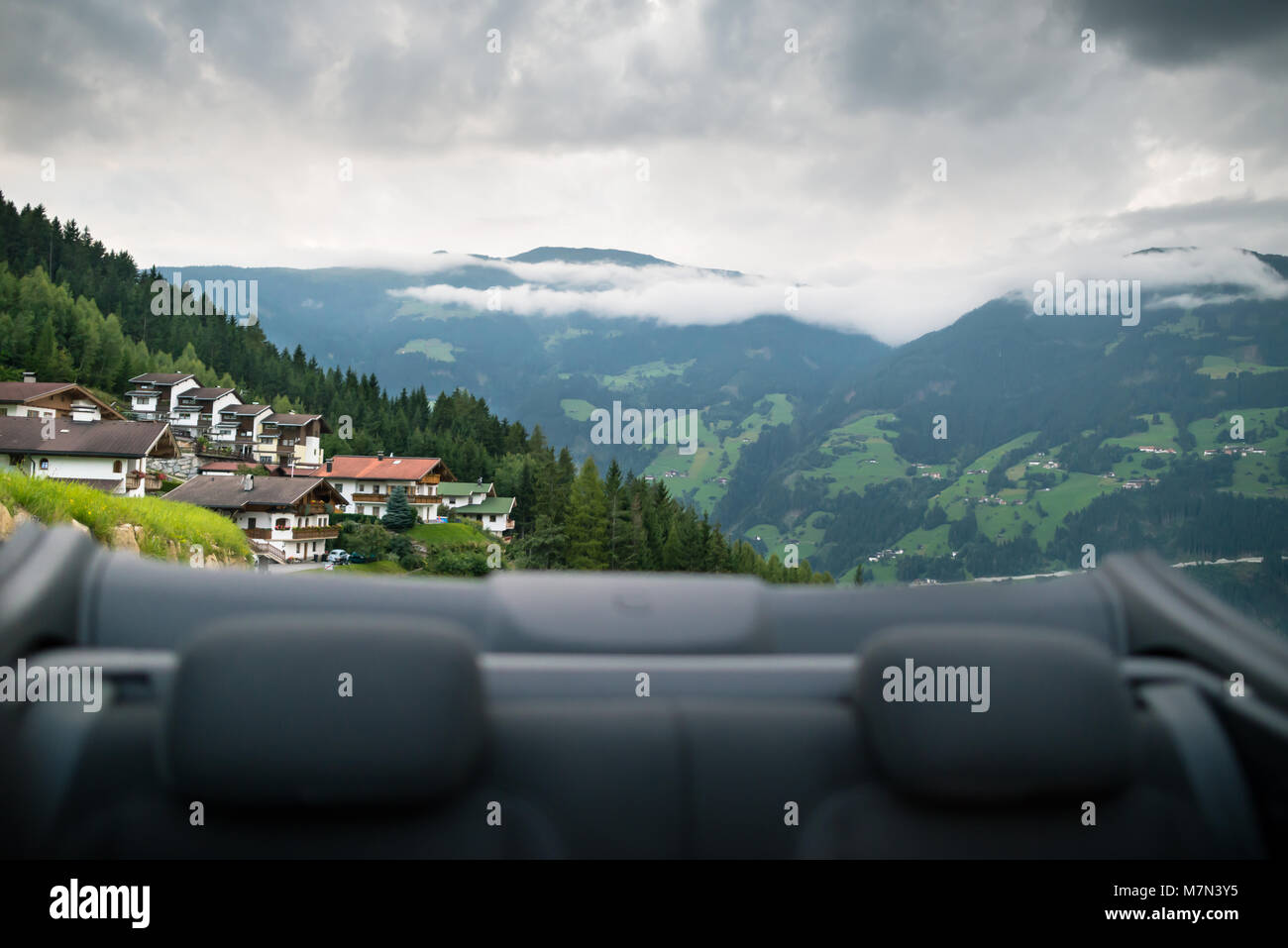 Road trip. Scenic back view from the cabriolet car on the Alp mountains and traditional buildings. Beautiful nature with green forest and hills. Stock Photo