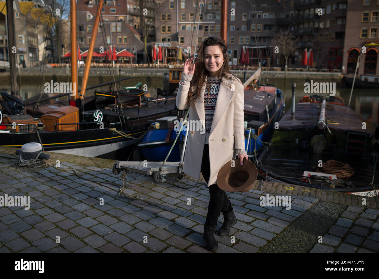 Young beautiful woman is smiling and standing near the old marina in Rotterdam, Netherlands. Elegant female tourist explore city in sunny day. Stock Photo