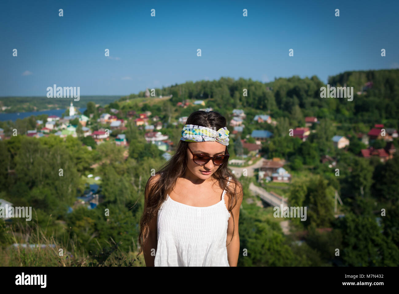 Young woman in sunglass and bandage stands on hill in summer day and looking down. Beautiful gir in the park on background of small village Stock Photo