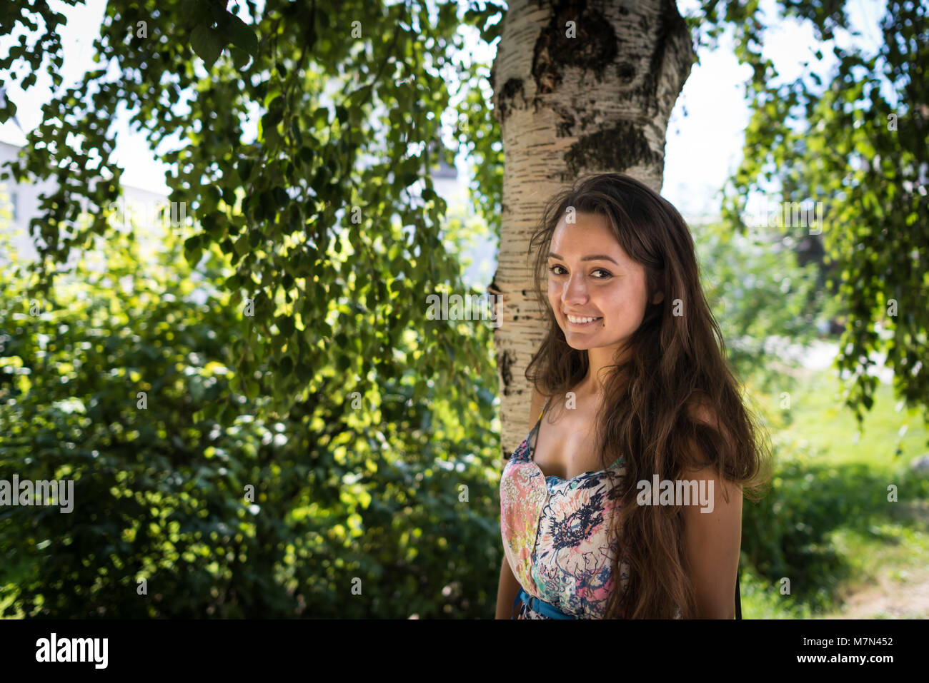 Young woman stands near tree in park on background of green leaves with sun rays. Cheerful girl is smiling under birch in warm summer day Stock Photo