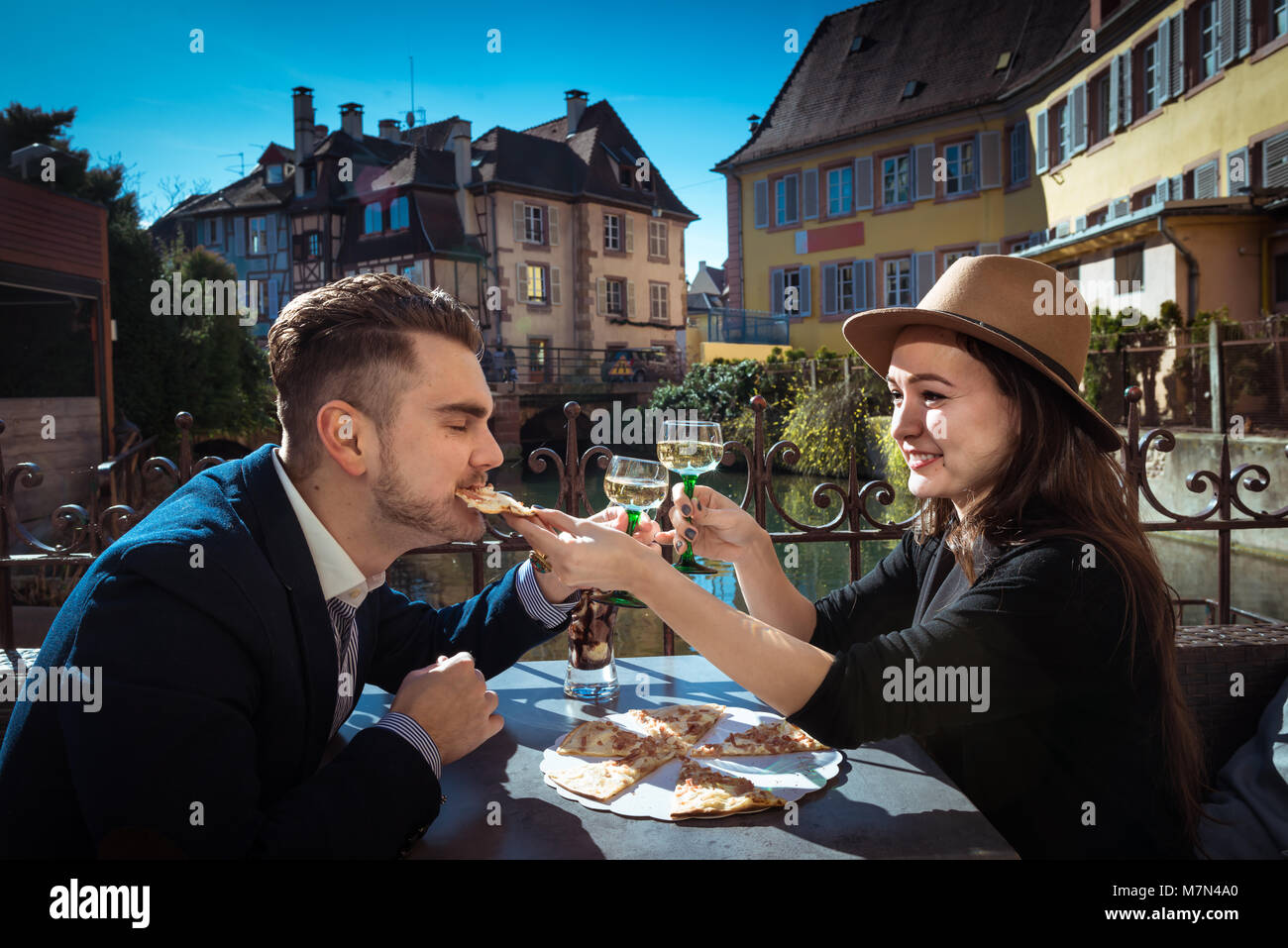 Young woman feeding boyfriend. Two stylish people sits together in local cafe with food and wine. Man in the suit eats pizza with girlfriend in Alsace Stock Photo