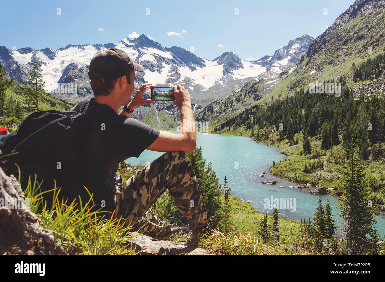 Tourist in black t-shirt takes photos with smart phone on peak of rock. Dreamy hilly landscape below. The beautiful landscape of the Altai Mountains.  Stock Photo