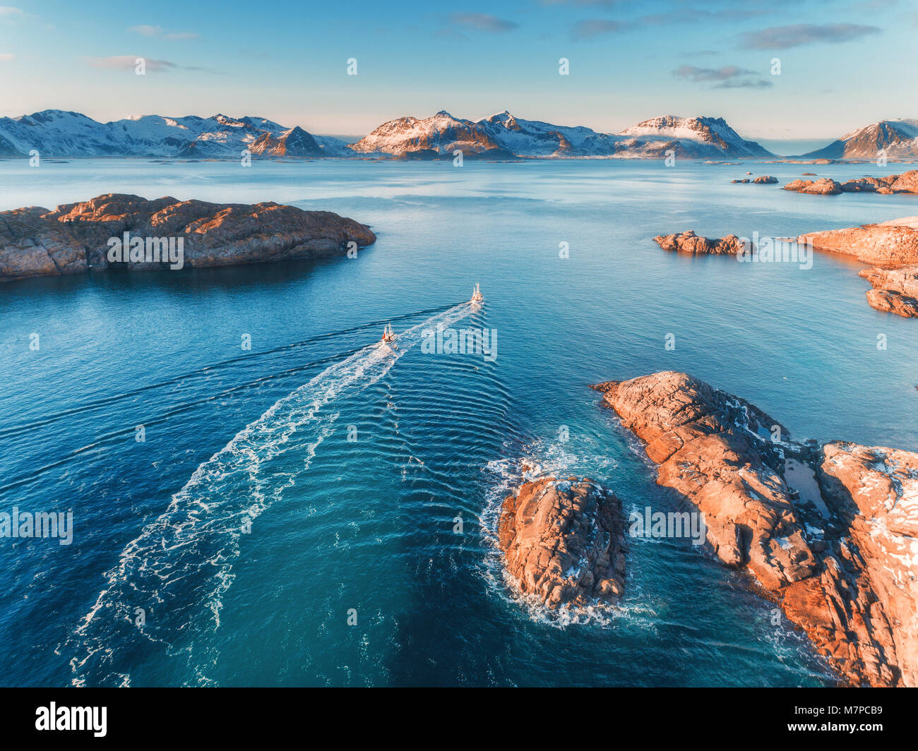 Aerial view of fishing boats, rocks in the blue sea, snowy mountains and colorful sky with clouds at sunset in winter in Lofoten islands, Norway, Land Stock Photo