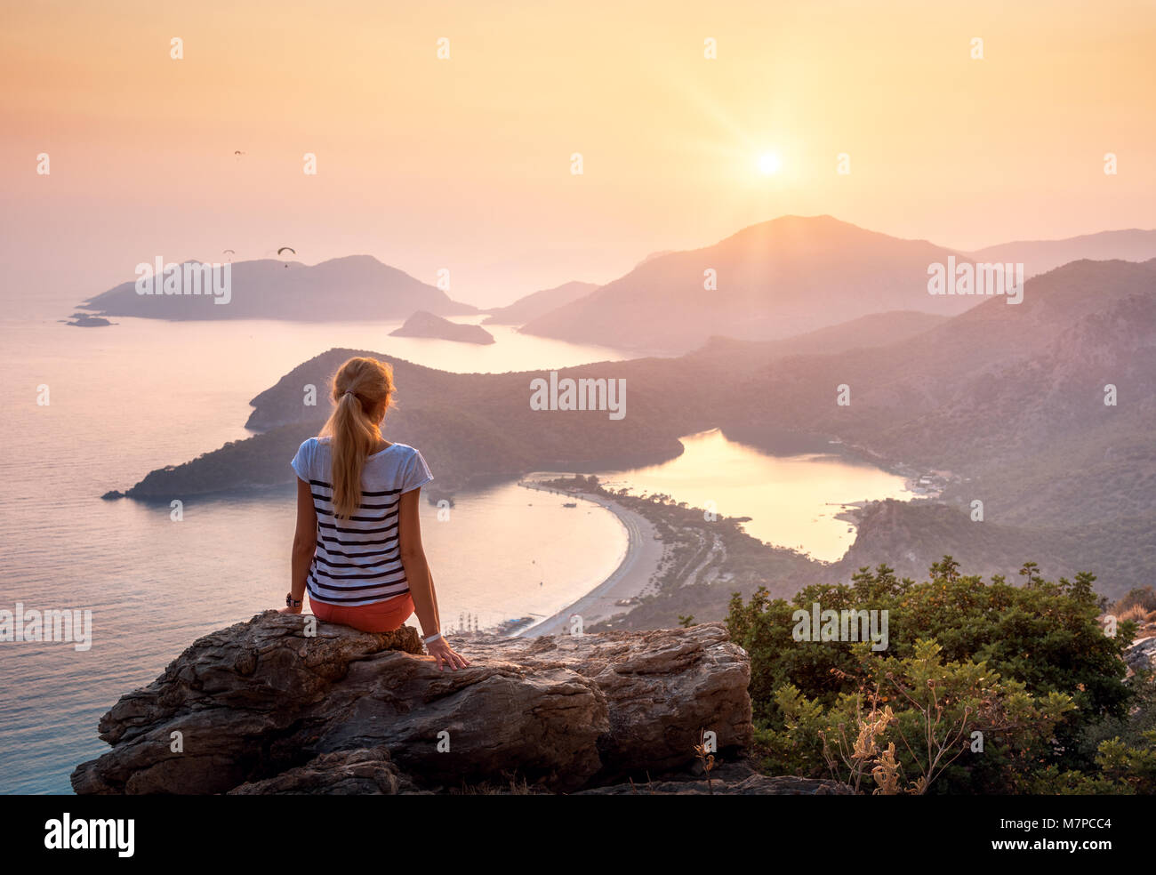 Young woman sitting on the top of rock and looking at the seashore and mountains at colorful sunset in summer. Landscape with girl, sea, mountain ridg Stock Photo