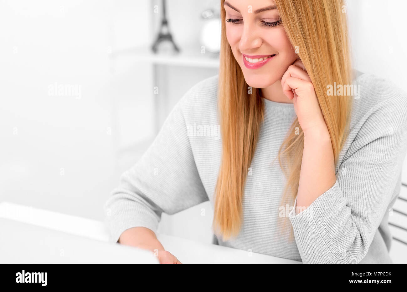 Young woman is working at a computer or laptop in a good mood. Blonde at her desk at home or in the office with a laptop and a red cup. Stock Photo