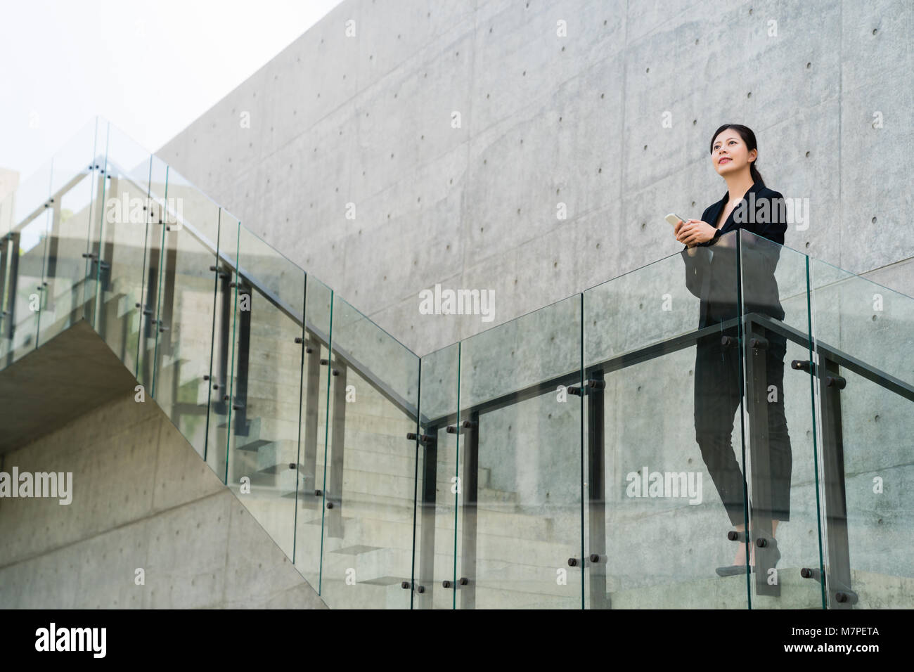 Pretty asian businesswoman stands at the business center stairs and stares thoughtfully into the distance, deliberating the state of affairs. Stock Photo