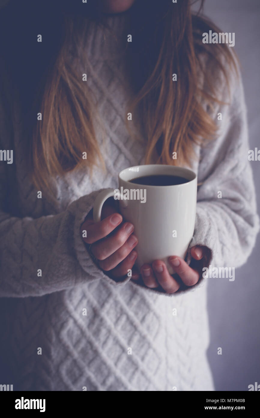 A midsection of a blonde haired girl holding up a white cup indoors and in the shadows. Stock Photo