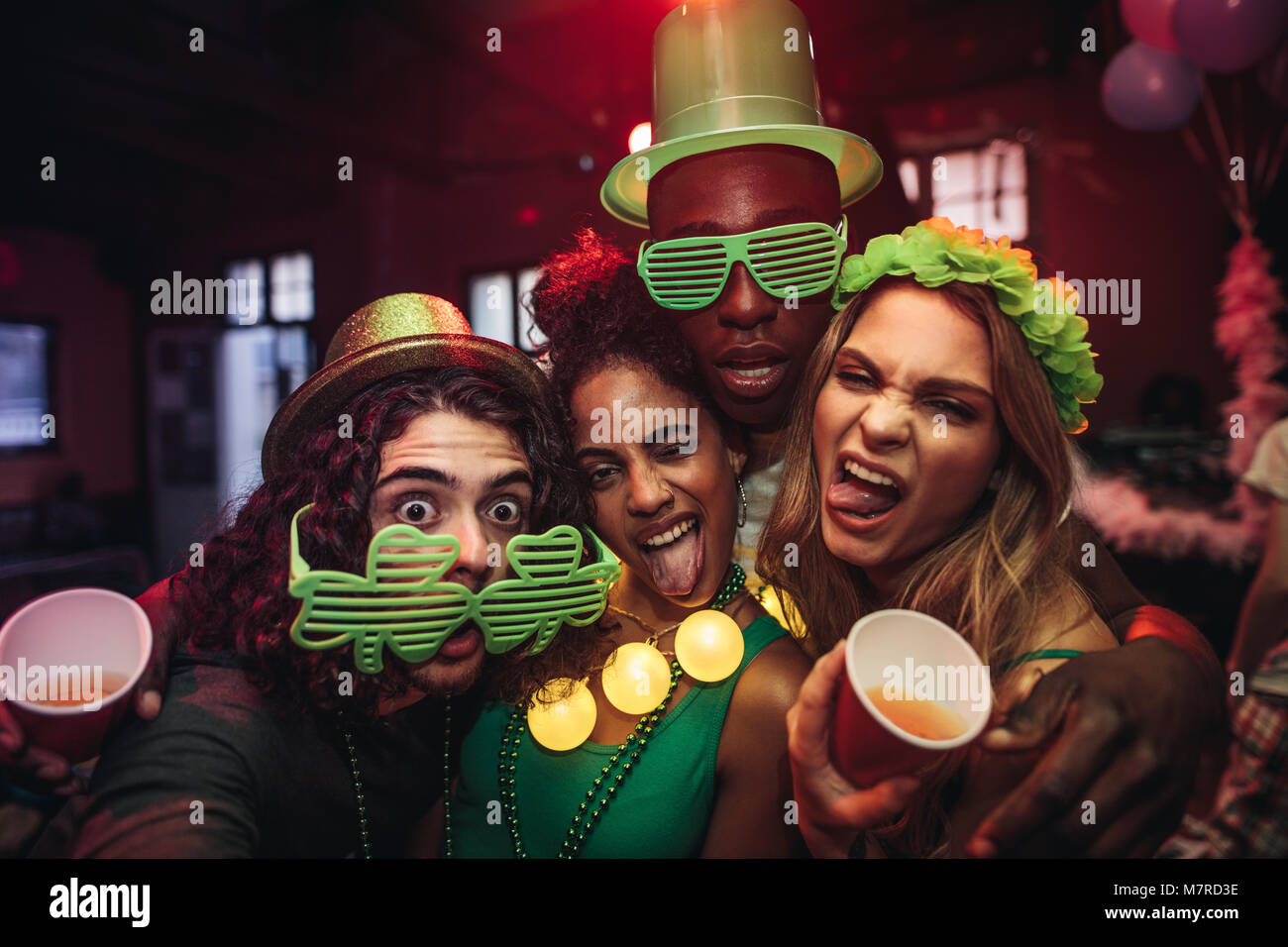 Crazy young friends enjoying a party at pub. Men and women in green clover party glasses and leprechaun hat at night club. People celebrating St.Patri Stock Photo