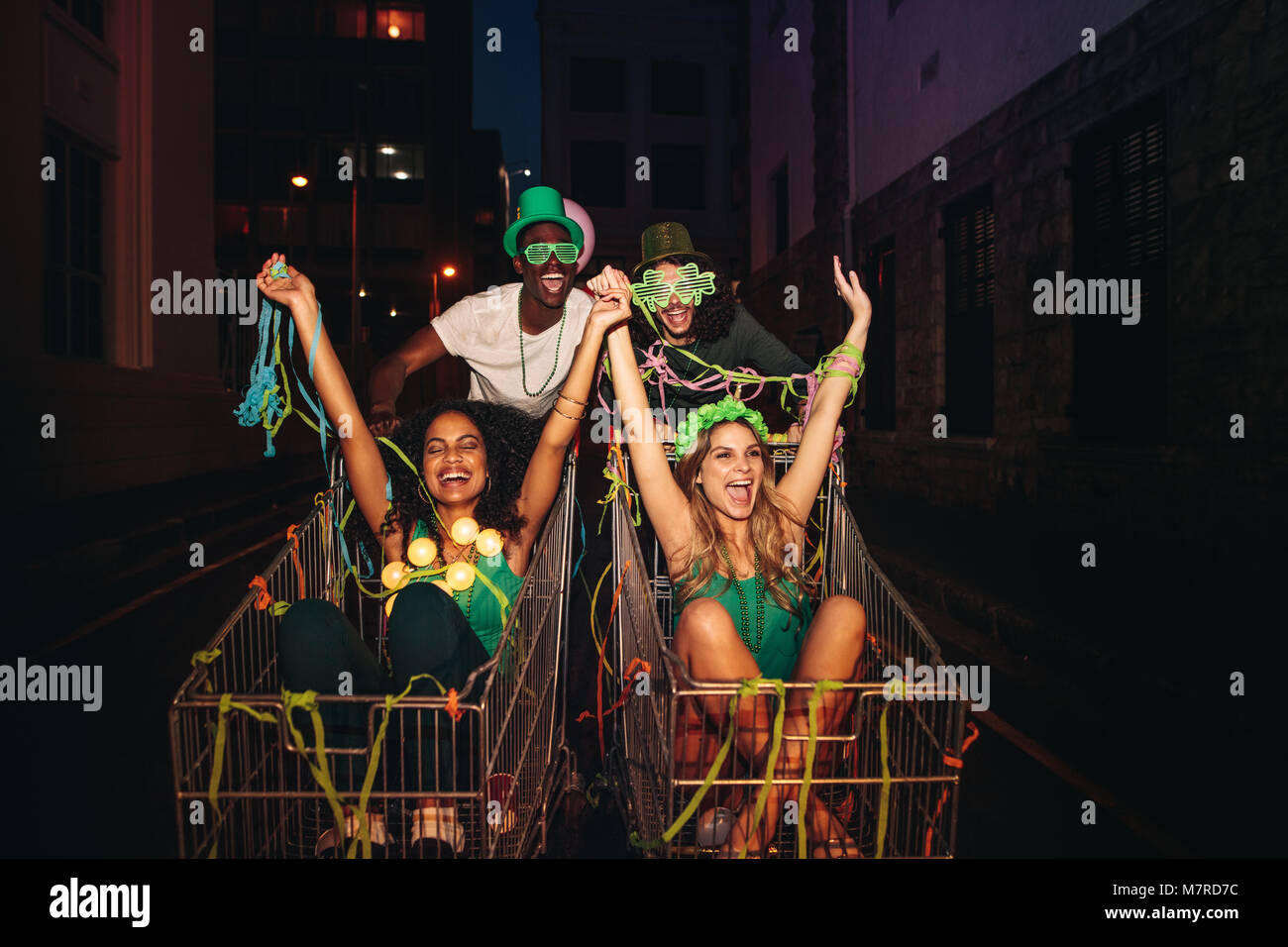 St.Patrick's day celebration on city street at night. Multi-ethnic people riding on shopping trolleys and having fun. Stock Photo