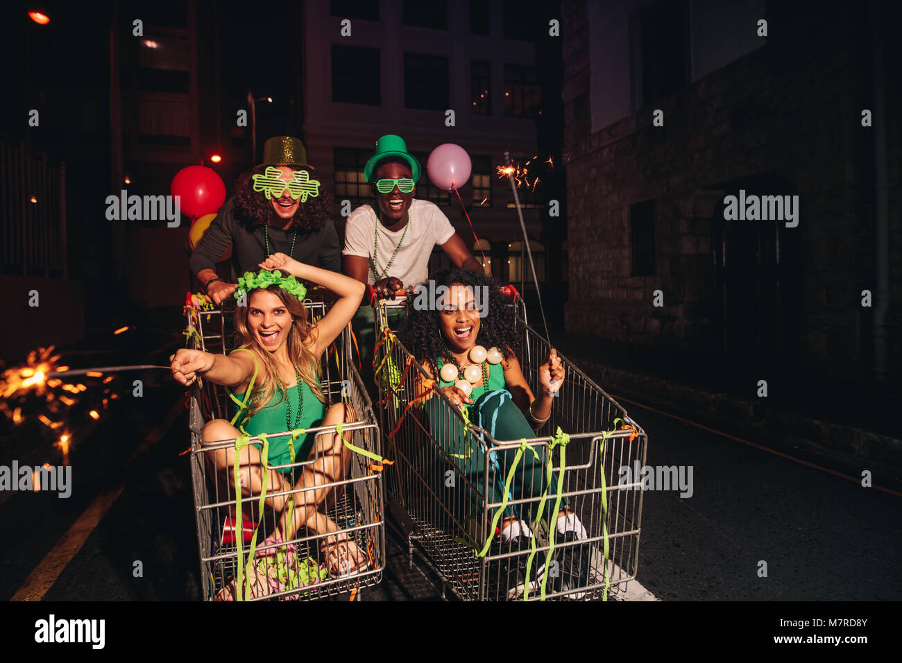 Men and women on shopping trolleys at night with sparklers on city street. Group of friends having fun on St.Patrick's day celebration at night. Stock Photo