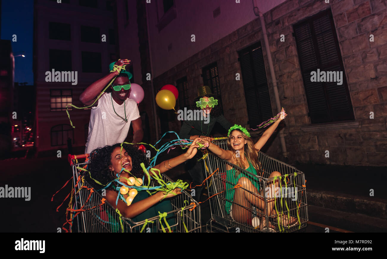 Young friends having fun on a shopping carts at night with confetti and balloons. Young people enjoying St.Patrick's day on city street at night. Stock Photo