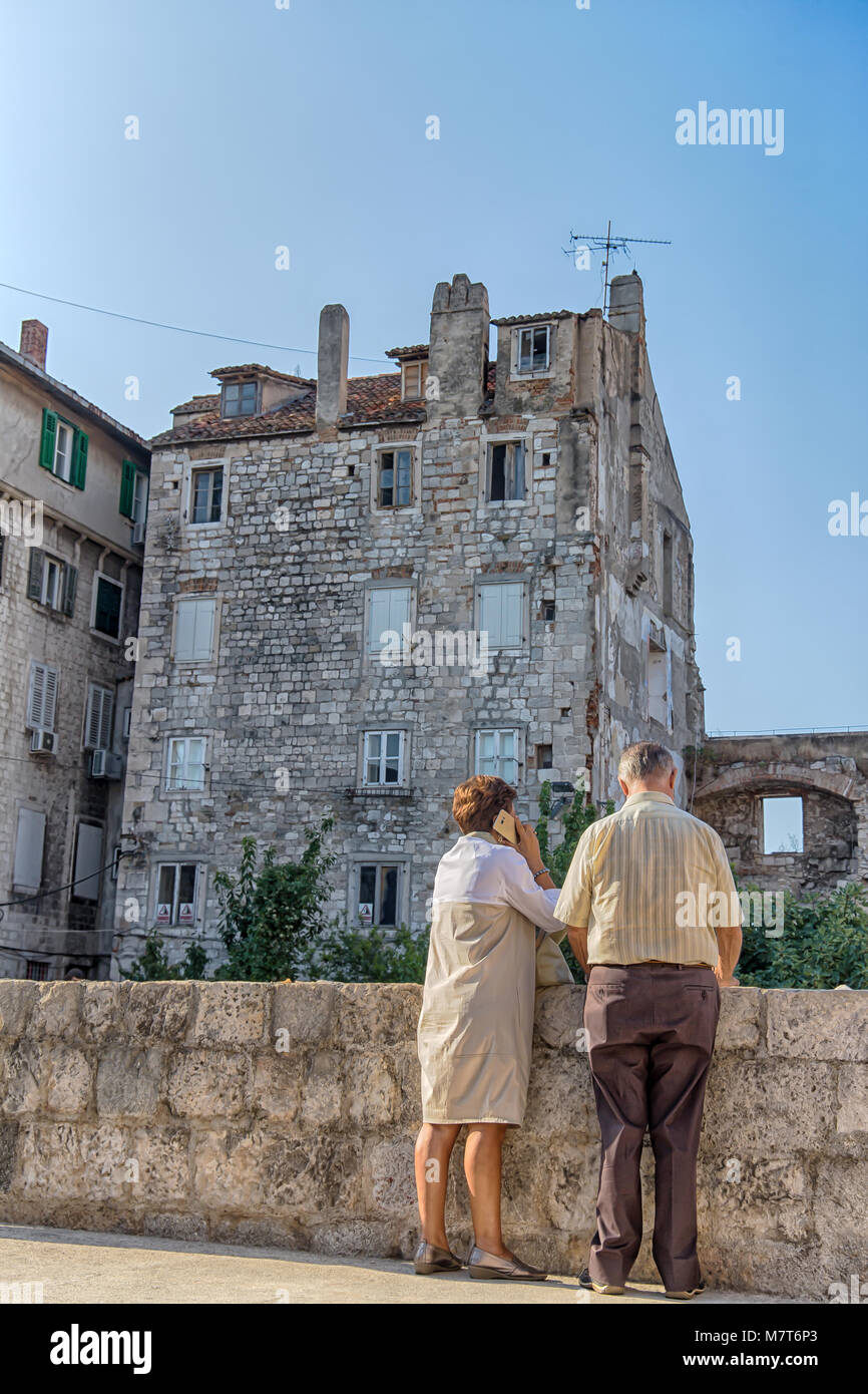 A twosome of elder people showing their backs, The woman speaking on the phone near a wall in front of a building Stock Photo