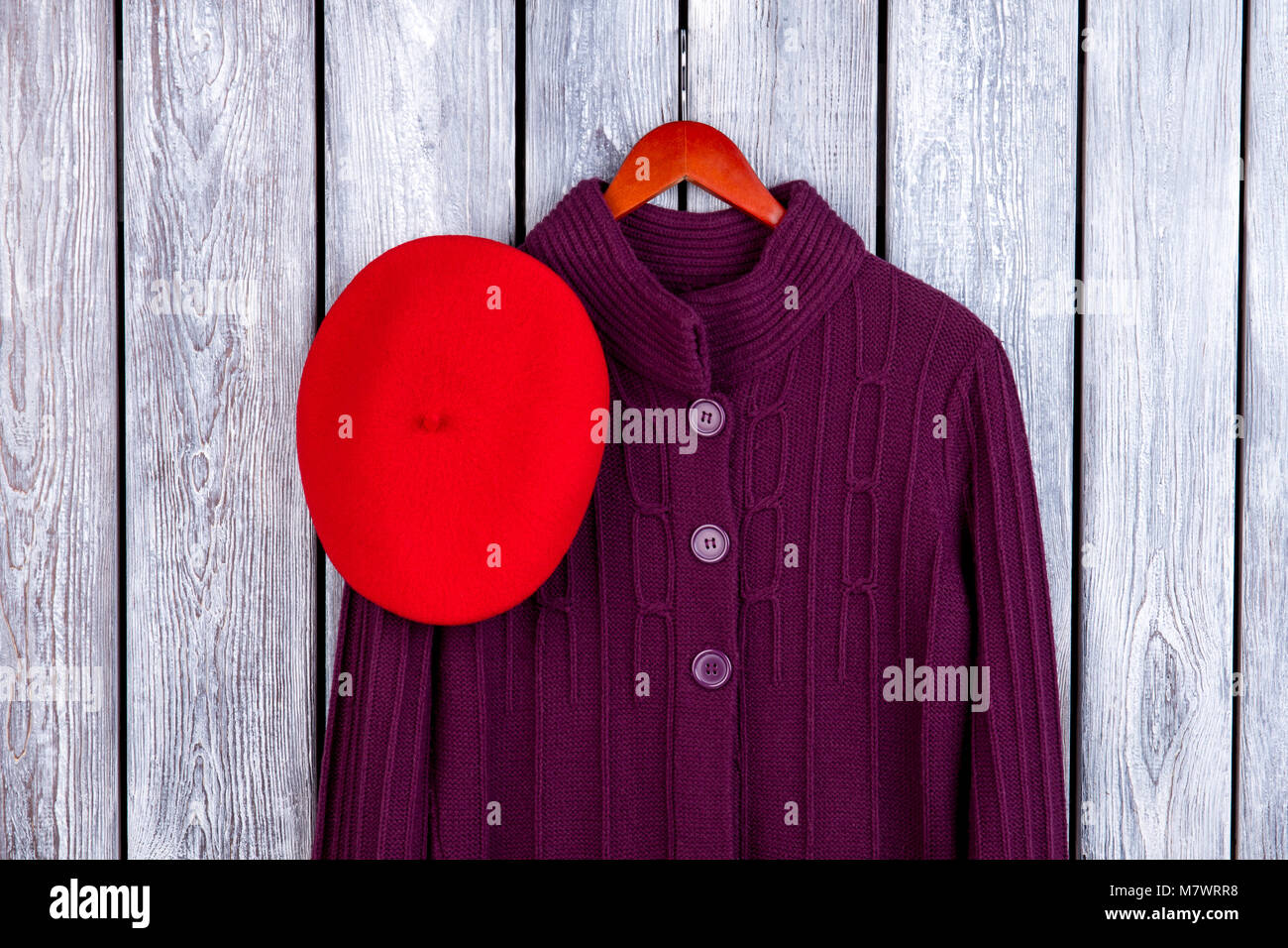 Winter women's coat and hat. Top view wool knitted coat on hanger and red beret. Dark wooden desk surface background. Stock Photo