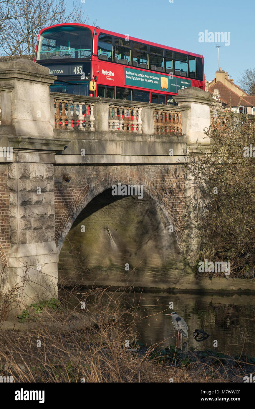 A crane (bird) on the river Brent as a red bus passes over a bridge near Hanwell in west London. Photo date: Sunday, February 25, 2018. Photo: Alamy Stock Photo