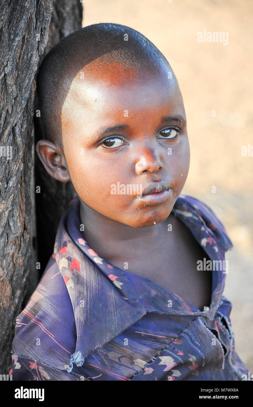 Unidentified young boy leans against a tree trunk in his local village. There is still much curiosity surrounding foreign visitors in Africa Stock Photo