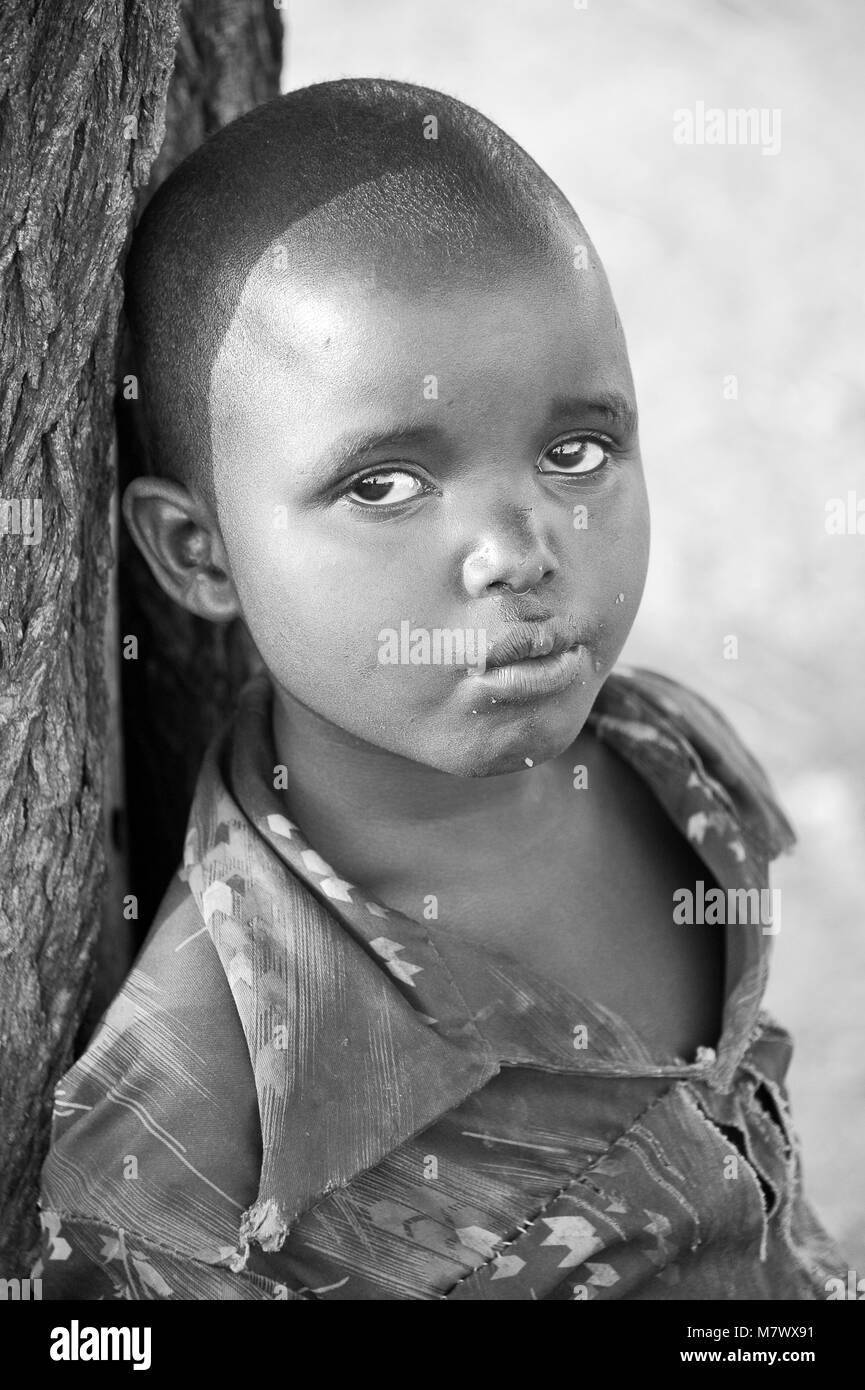 Monochrome portrait of a young boy leaning against a tree trunk in his local village. There is still much curiosity surrounding foreigners in Africa Stock Photo