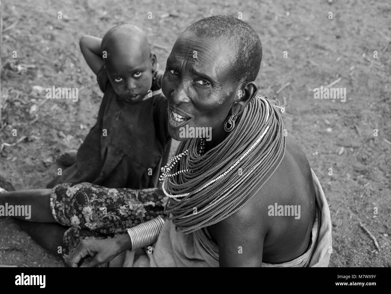 Monochrome portrait of an unidentified mother and child, wearing traditional ornamental necklace of red beads, sits outside hut in local village. Stock Photo