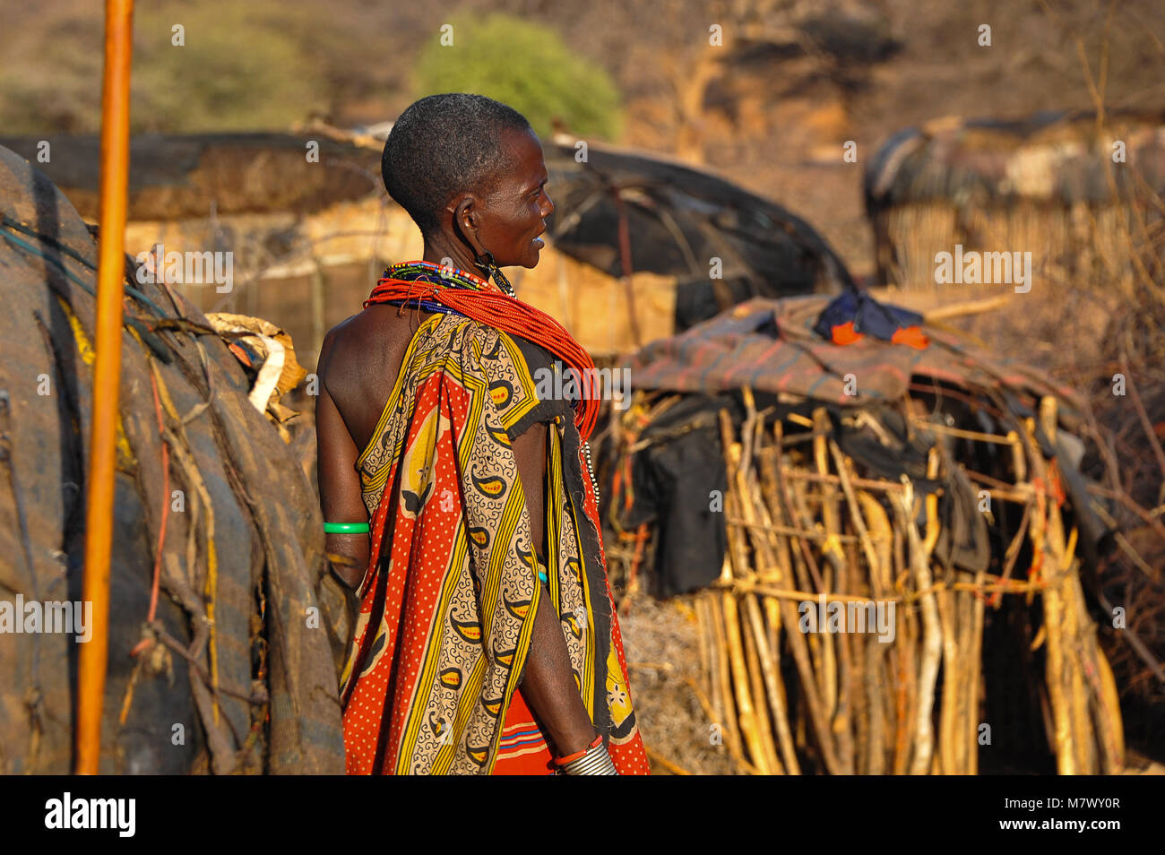 African tribeswoman wearing colourful dress stands with back to camera in traditional village or kraal. Portrait showing woman wearing ornate jewellry Stock Photo