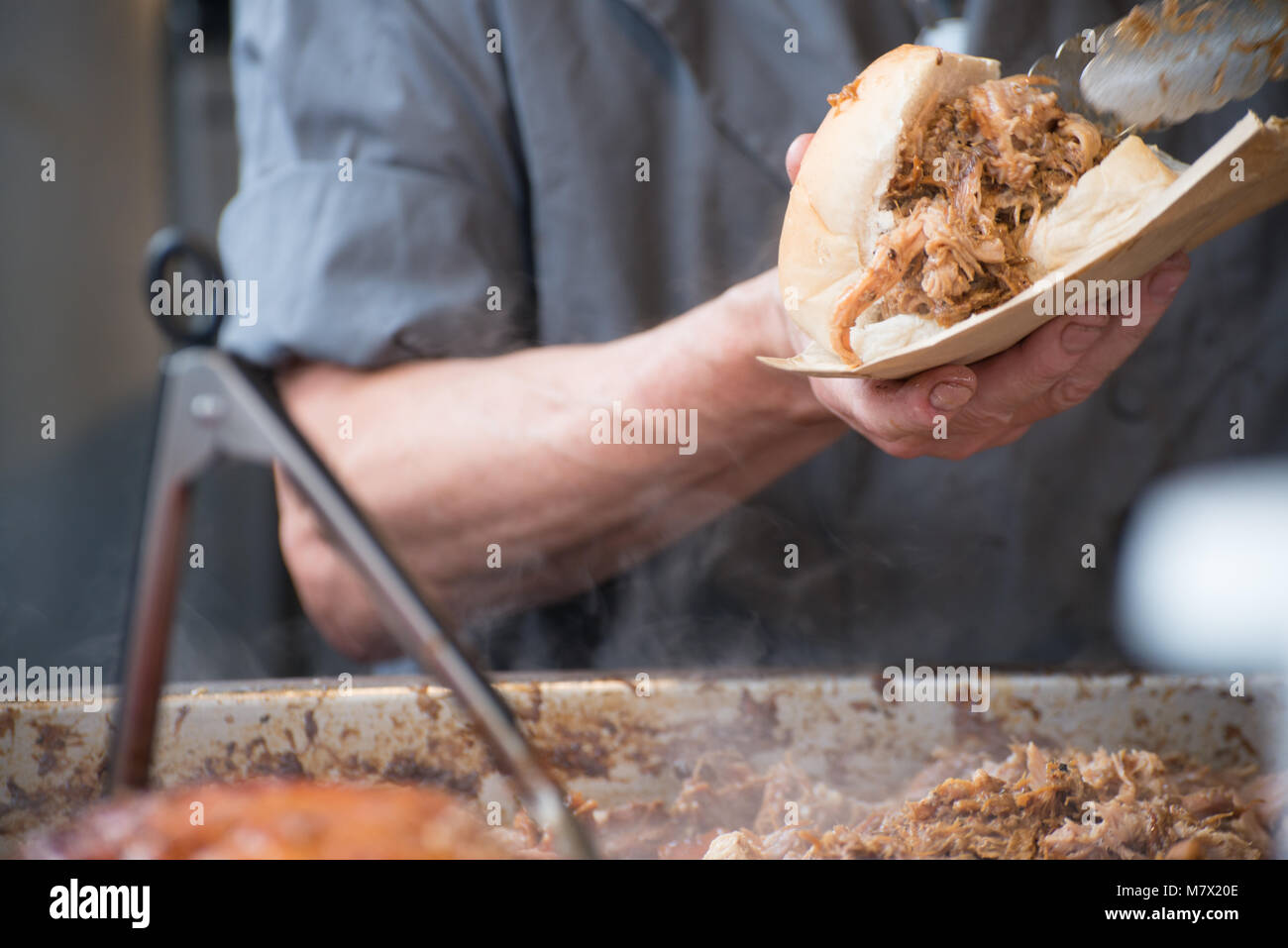 Man in grey shirt serving pulled pork street food holding sandwich with rind and serving tongs in the foreground Stock Photo