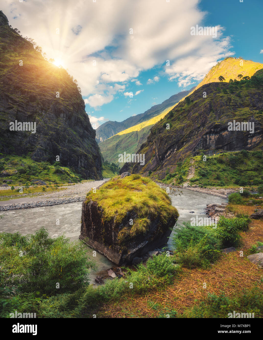 Amazing scene with mountains, beautiful river, green forest, blue sky with clouds and big stone in water in autumn in Nepal at sunset. Mountains at br Stock Photo