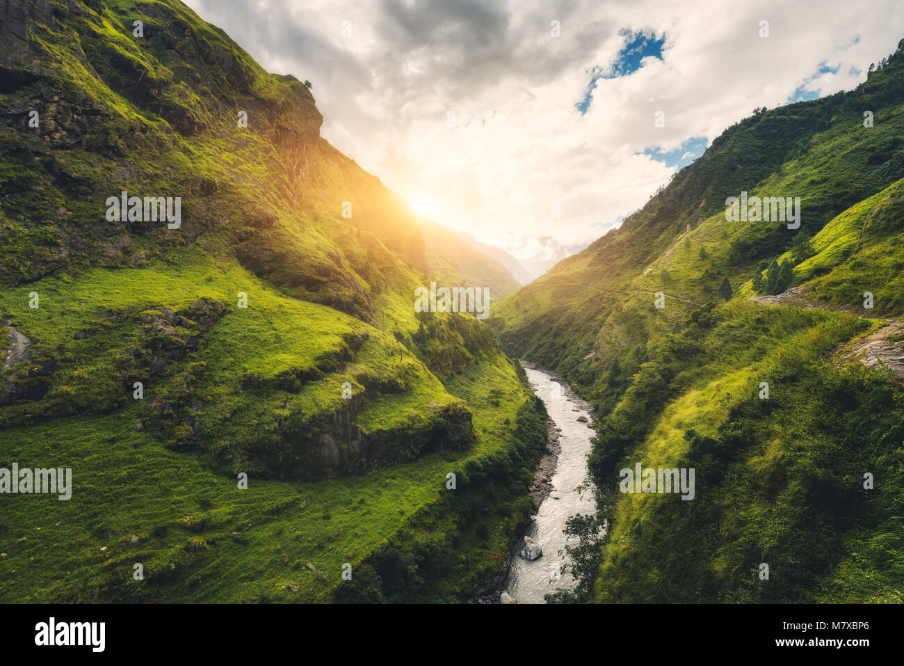 View with amazing mountains covered green grass, river, meadows and forest, blue sky with clouds, sun in autumn in Nepal at sunset. Mountain valley at Stock Photo