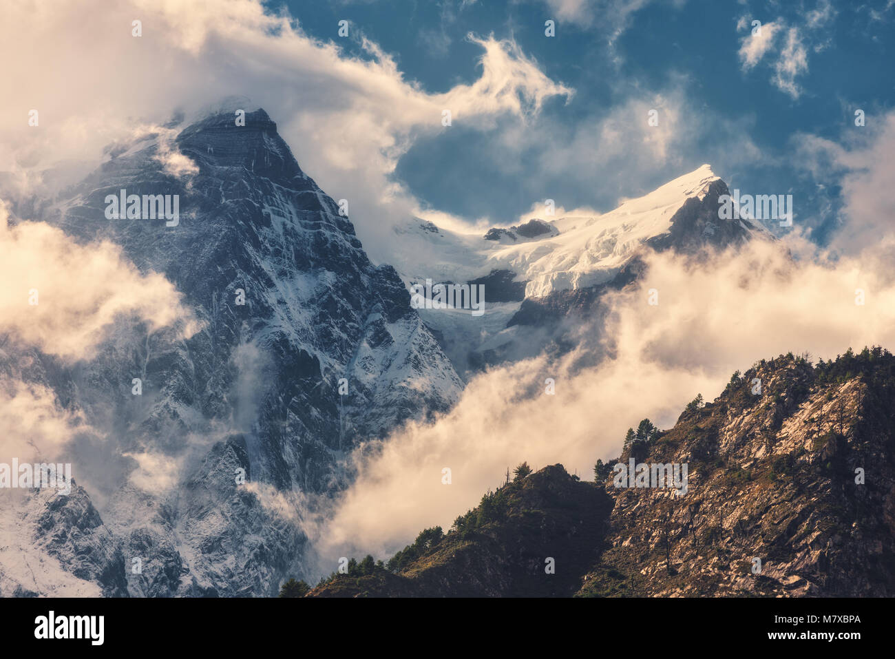 High mountains with snowy peaks in clouds at bright sunny evening in Nepal. Colorful landscape with beautiful rocks and dramatic cloudy sky at sunset. Stock Photo