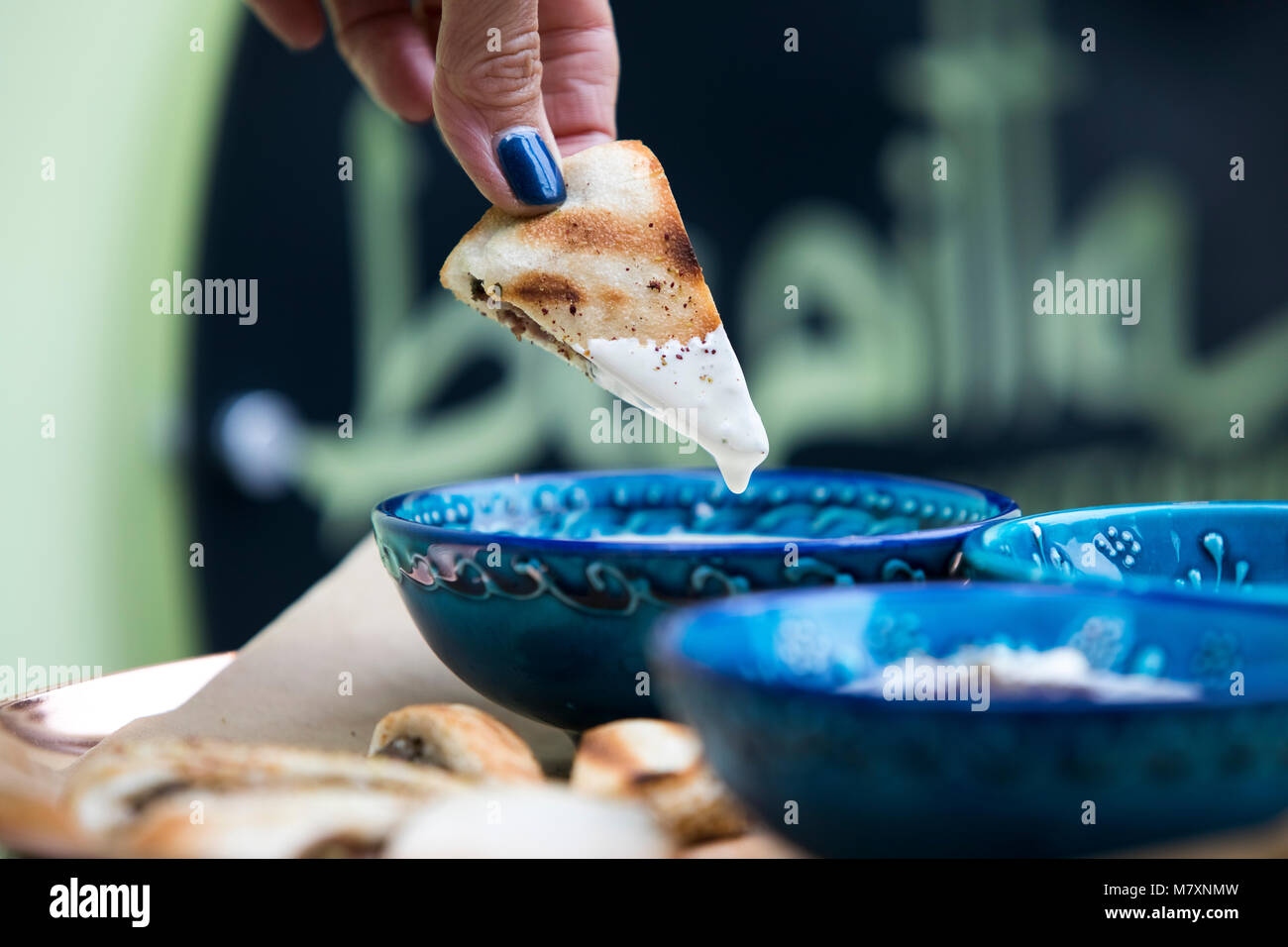 Meat stuffed flatbread being dipped in tahini dip held by womans hand with blue nails.  Blue bowl.  Middle Eastern. Stock Photo